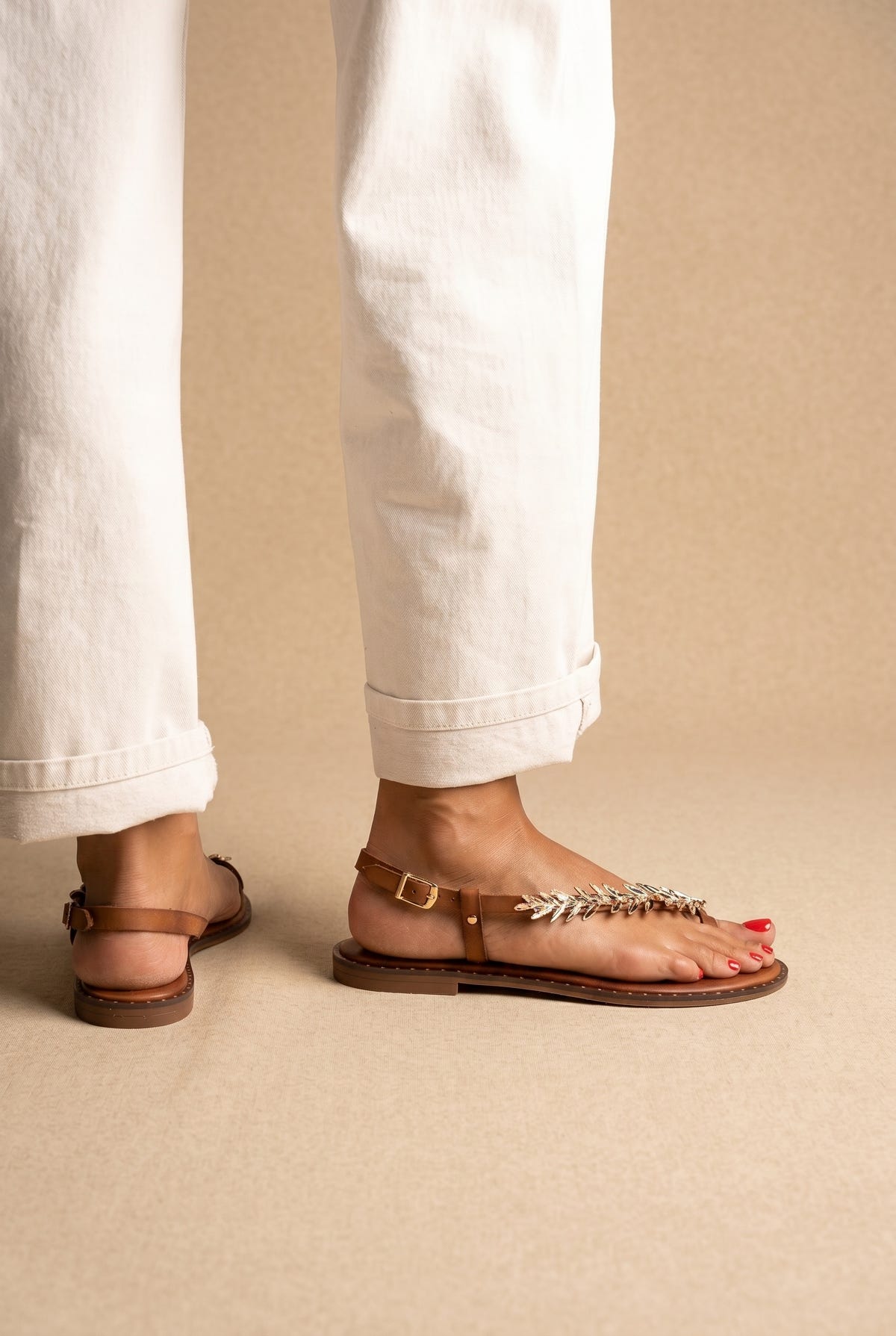 Tan leather thong sandals with gold leaf-shaped toe strap and ankle buckle, worn on a model with white trousers, side view