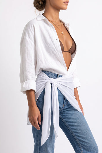 Model wearing white sheer mini pareo tied over open white linen shirt, brown bikini top and blue jeans