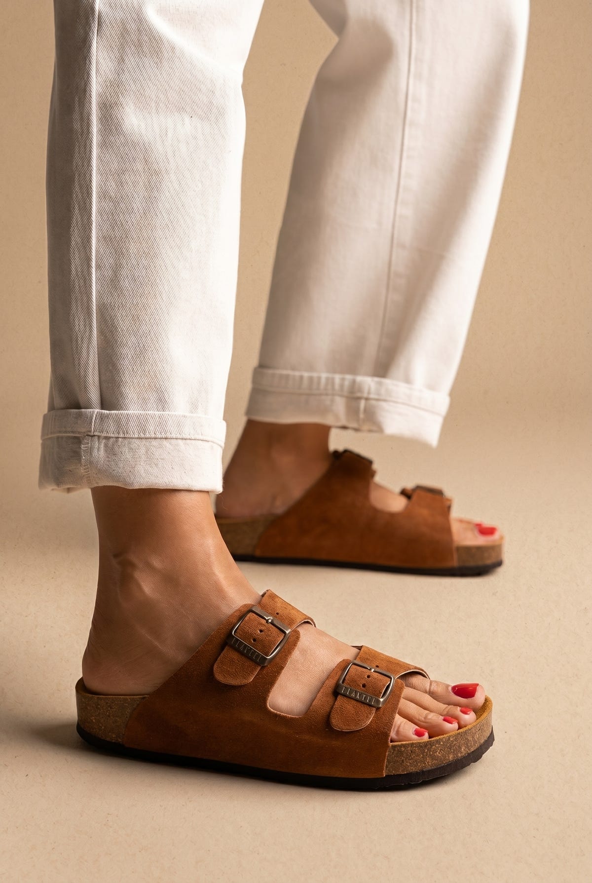 Tan suede two-buckle cork footbed slides with dark sole, side view worn on a model with white cuffed trousers