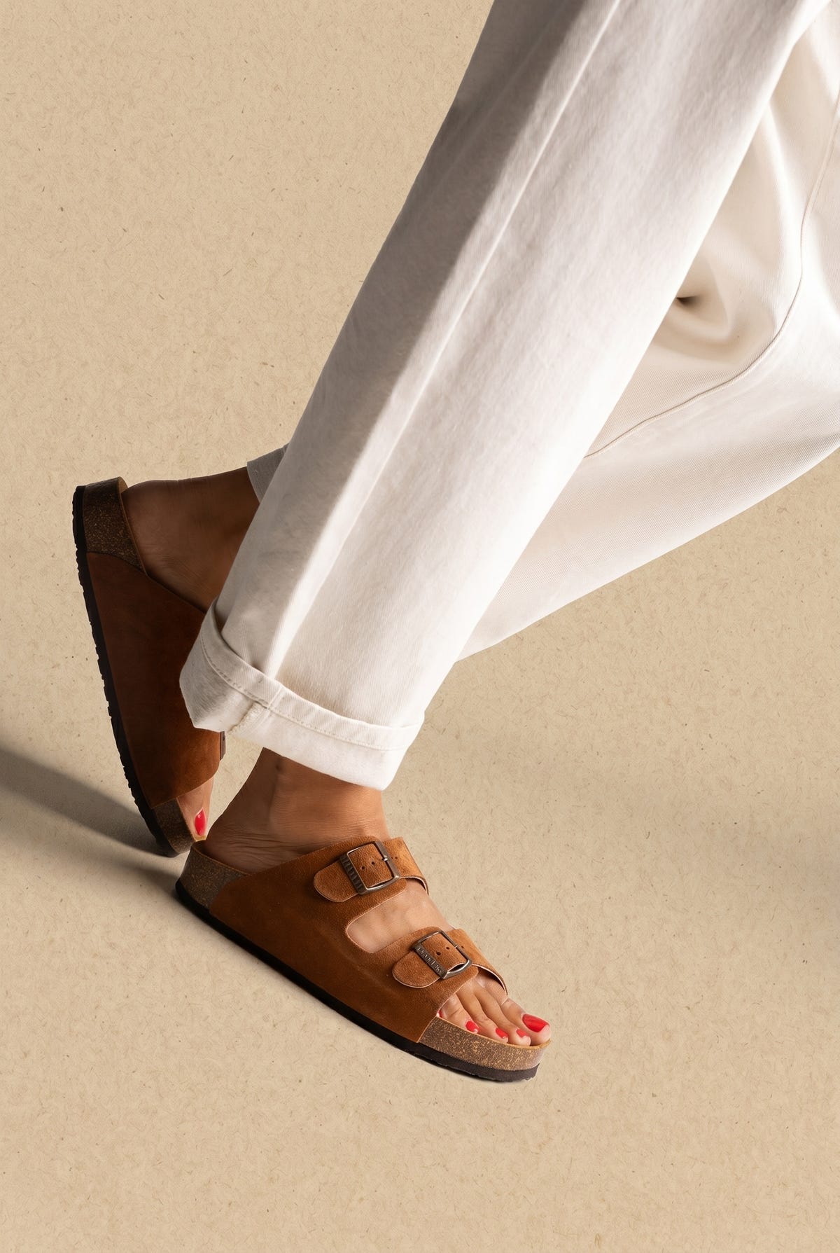 Tan suede two-strap slides with metal buckles and cork footbed, worn on a model with white trousers, three-quarter view