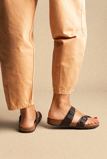Dark brown buckled toe-loop slides with cork footbed and dark sole, worn on a model with tan trousers, side view