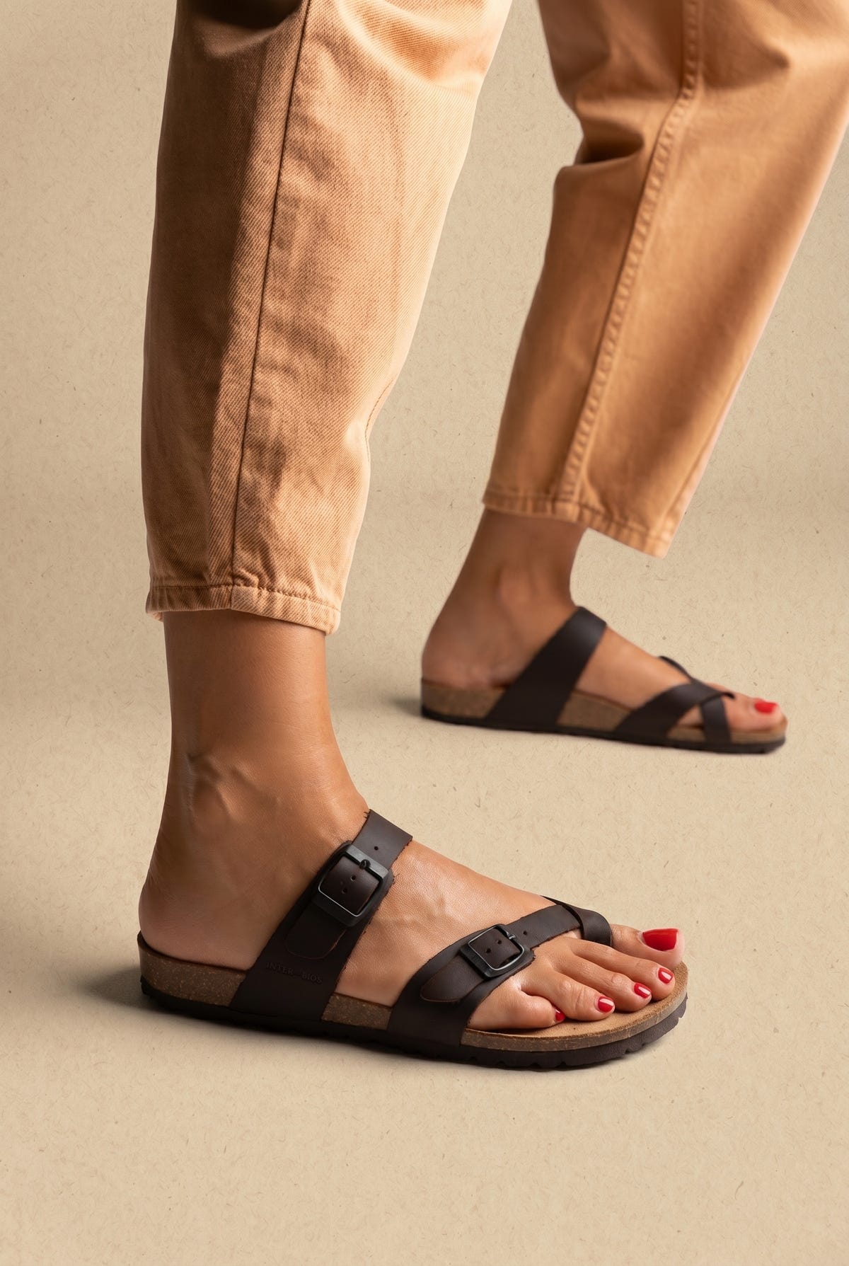 Dark brown toe-loop slides with two buckle straps and cork footbed, worn on a model with tan trousers, angled view
