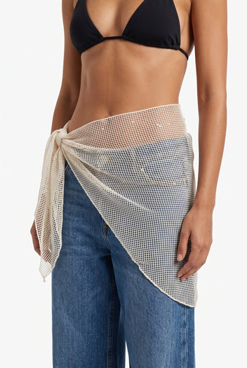 Front view of model wearing beige mesh pareo with scattered sequins tied at the hip over blue jeans and black bikini top