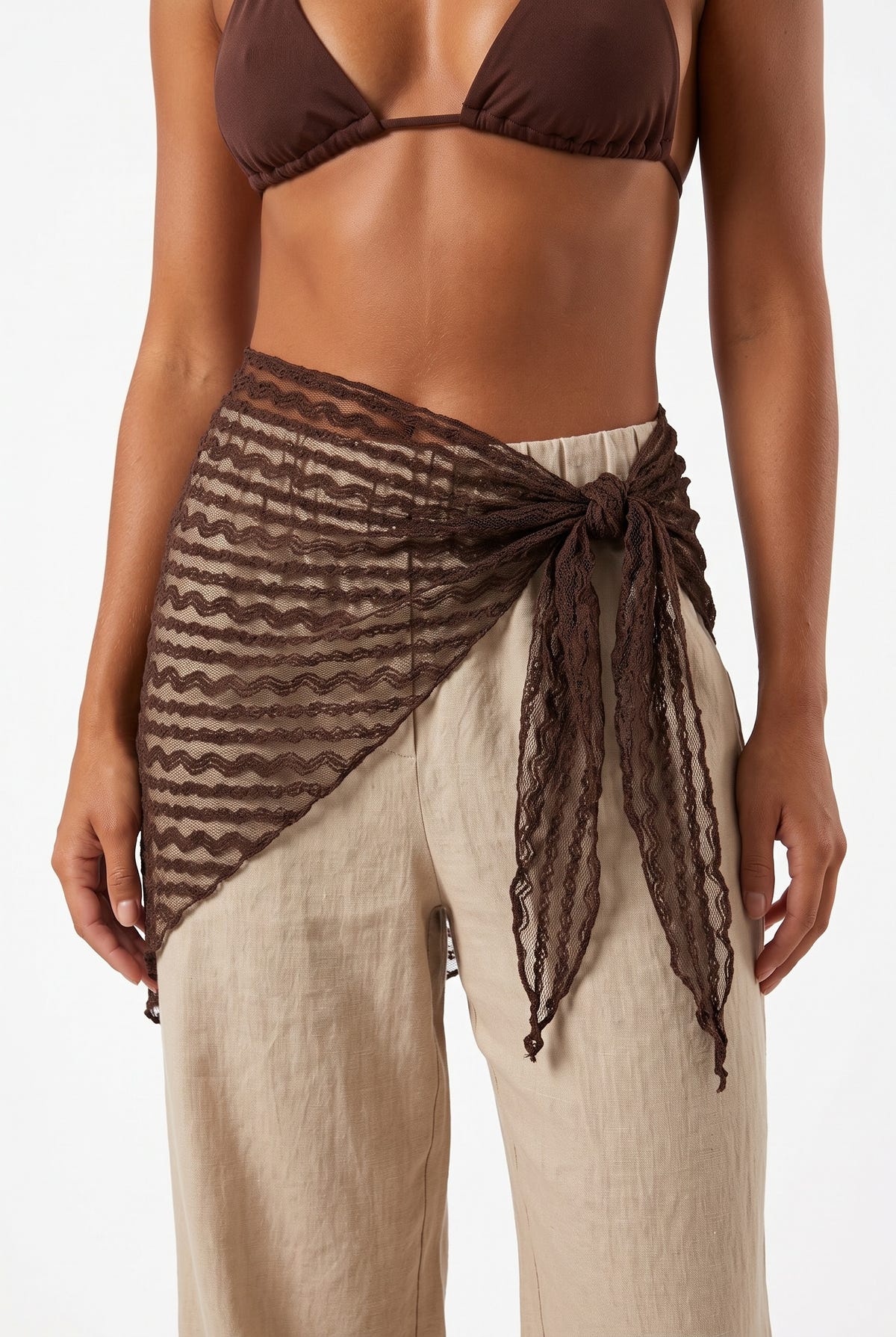 Front view of model wearing brown sheer lace sarong with side knot over beige linen trousers and brown bikini top
