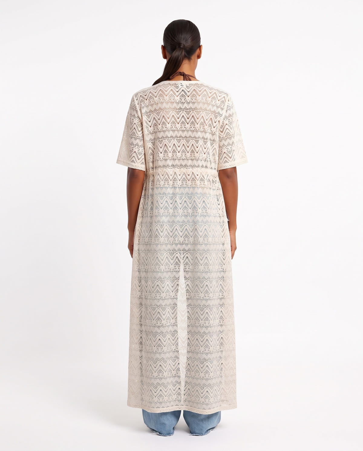 Rear view of model wearing long beige crochet lace kaftan with short sleeves over light blue jeans