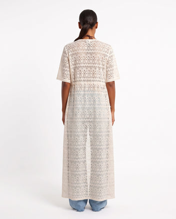 Rear view of model wearing long beige crochet lace kaftan with short sleeves over light blue jeans