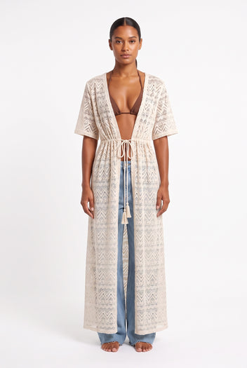 Front view of model wearing long beige crochet lace kaftan with tie waist, short sleeves and light blue jeans
