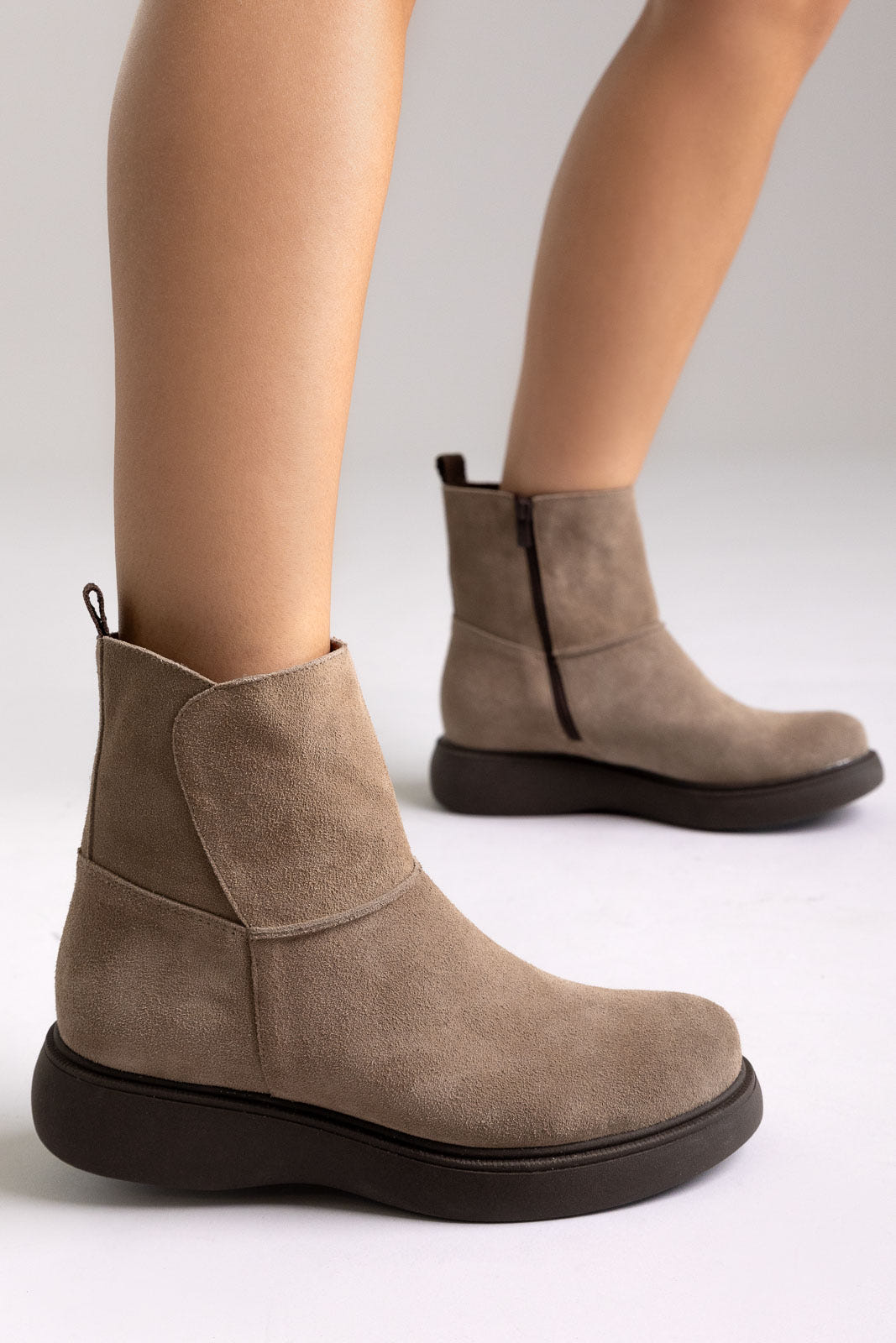 Taupe suede flat ankle boots worn on both feet, close side view showing zip detail