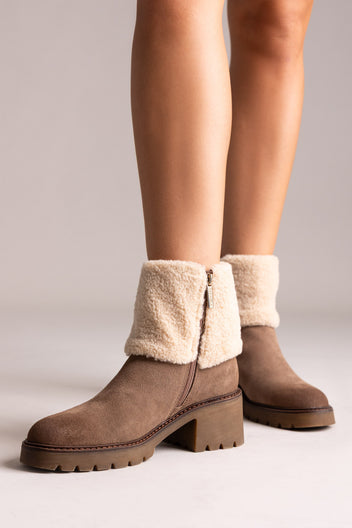 Model legs in taupe suede shearling-cuff ankle boots with block heels, front view on a beige backdrop