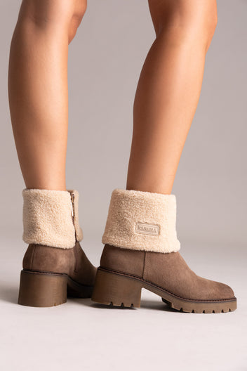 Model legs in taupe suede shearling-cuff ankle boots with block heels, front view on a light grey background