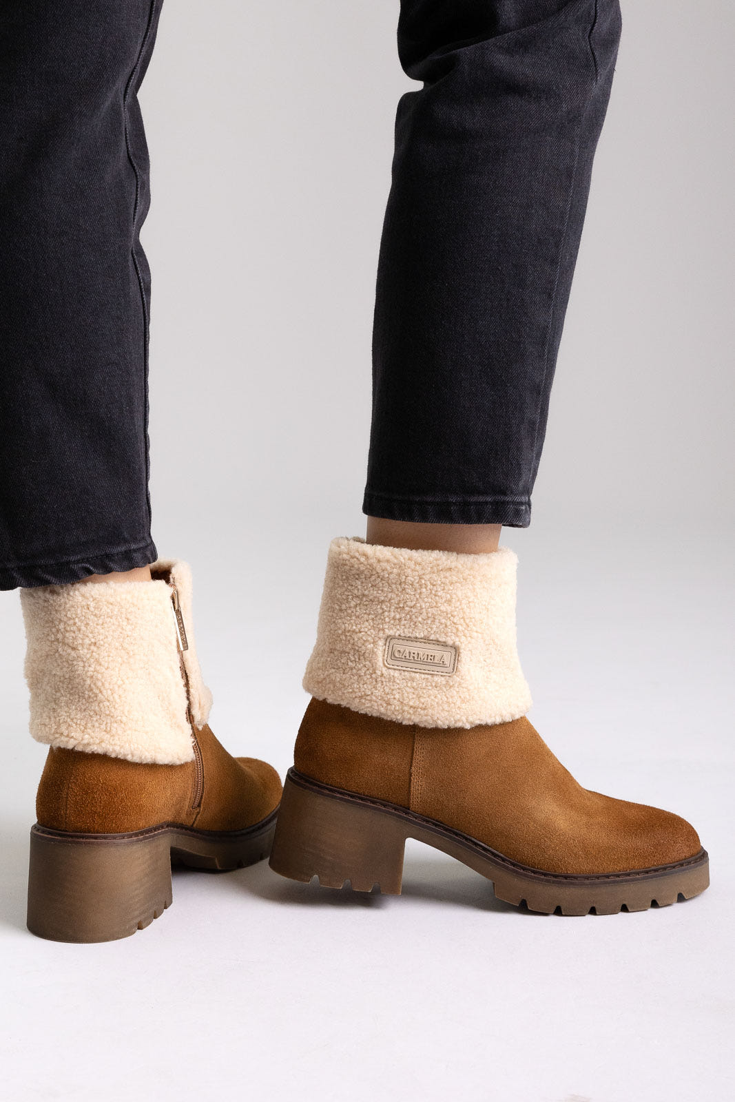 Model legs in tan suede shearling-cuff ankle boots with block heels, worn with dark jeans, angled view on light grey