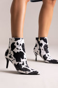 Black and white cow-print pointed-toe stiletto ankle boots with kitten heel, side view on a model