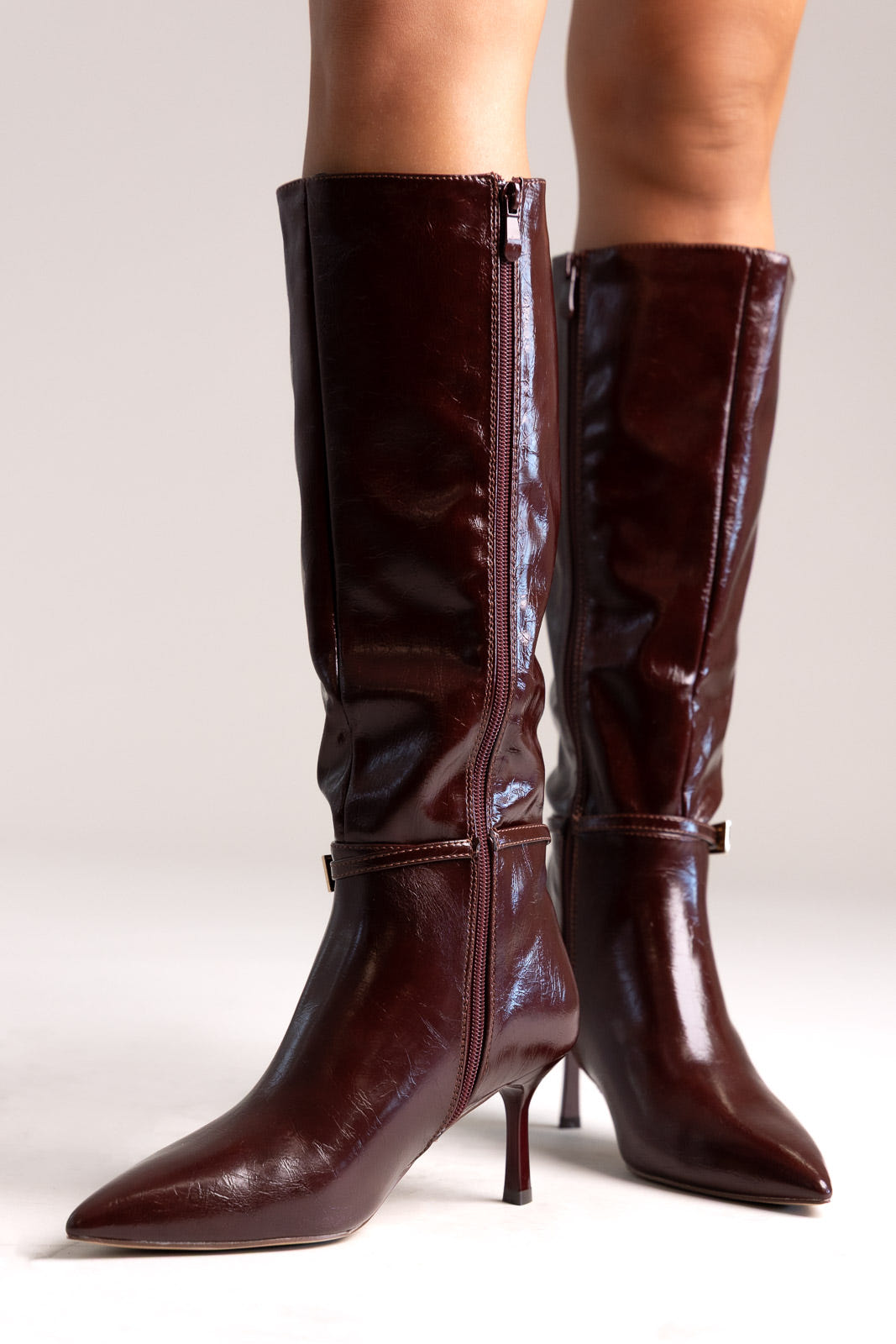 Dark burgundy patent faux leather knee-high boots with pointed toe, kitten heel and gold buckle strap, side view
