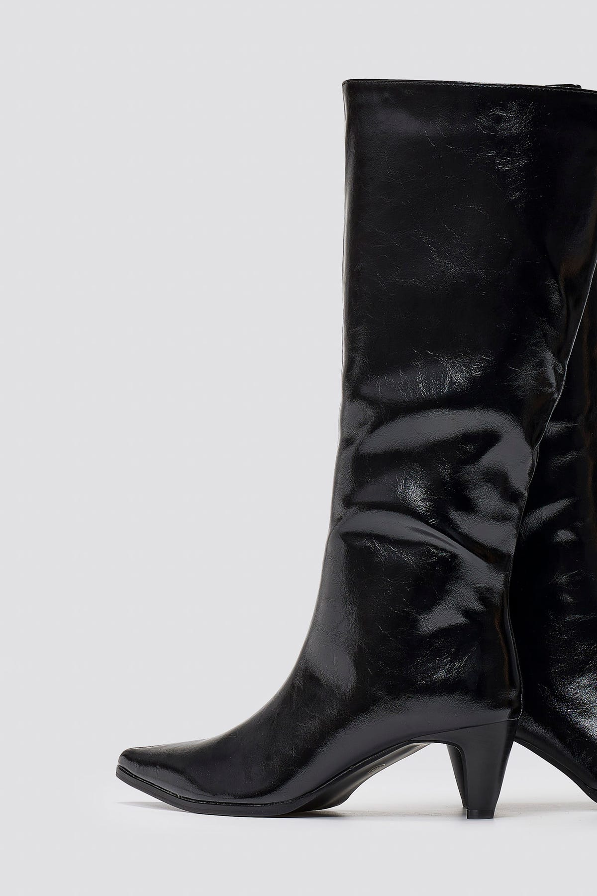 Black crinkled patent leather knee-high boots with pointed toe and cone heel, close-up of shaft on grey