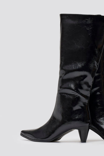 Black crinkled patent leather knee-high boots with pointed toe and cone heel, close-up of shaft on grey