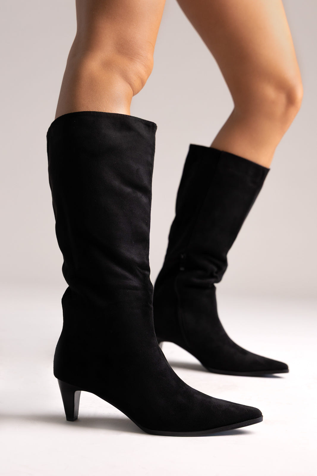 Black faux suede pointed-toe knee-high boots with kitten heel, side view on a model