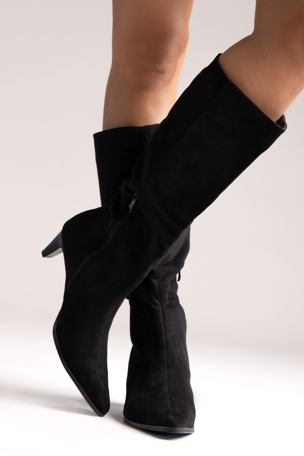 Black faux suede pointed-toe stiletto knee-high boots with slouchy shaft, angled side view on a model
