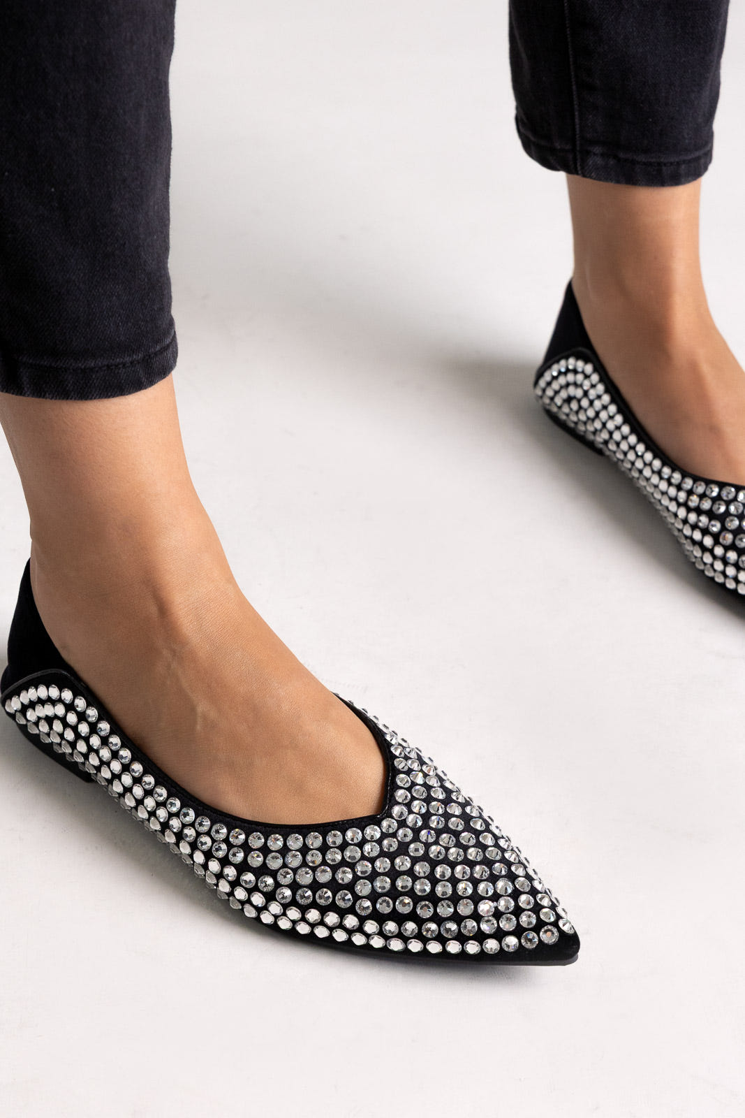 Black suede pointed-toe flats with crystal embellishment, close-up on a model in black jeans