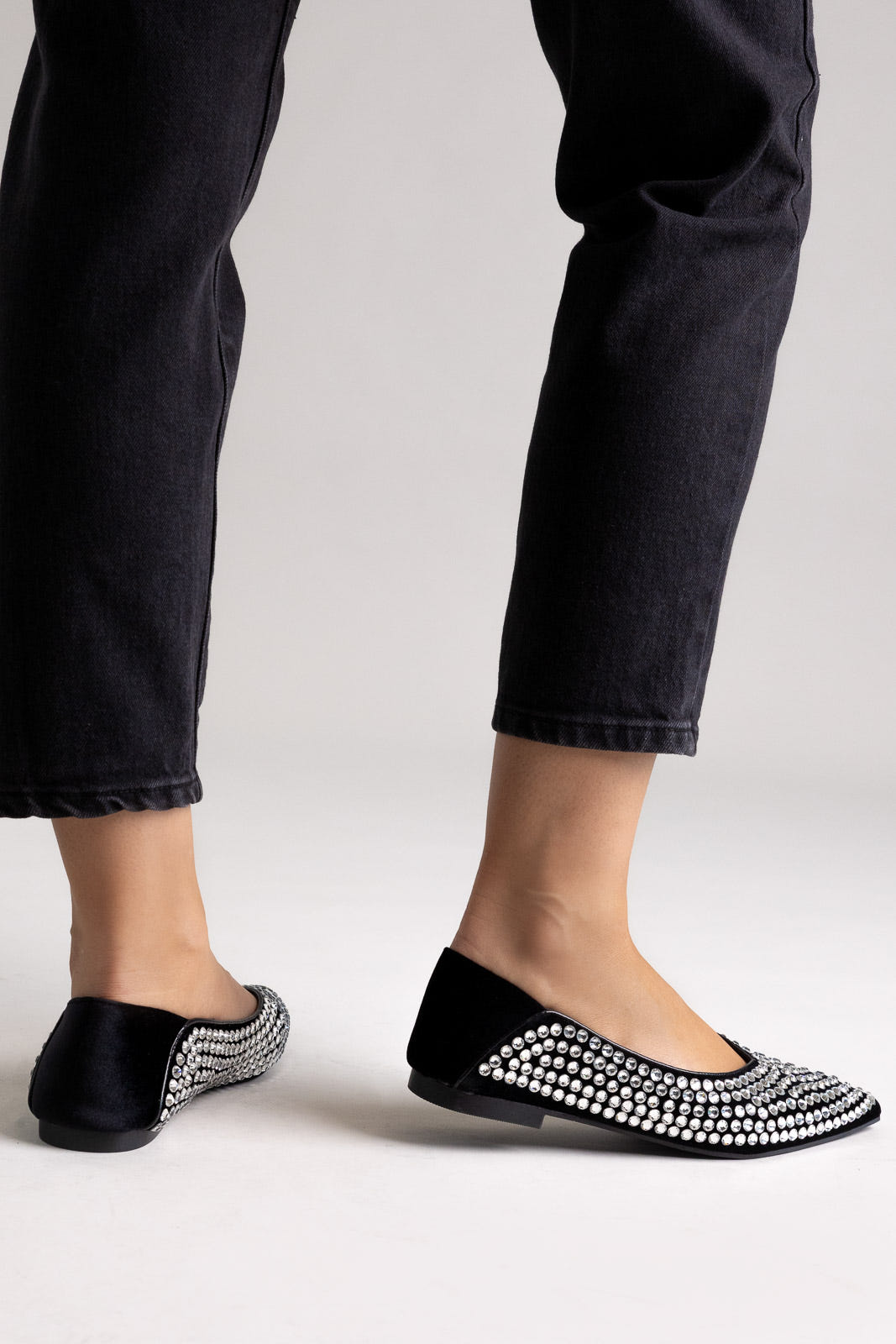 Black pointed-toe flats with crystal embellishment, angled view on a model wearing black jeans