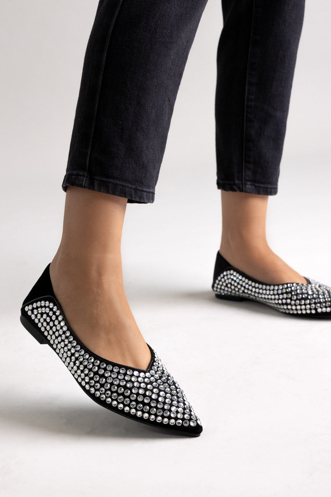 Black suede pointed-toe flats with rhinestone embellishment, angled view on a model in black jeans
