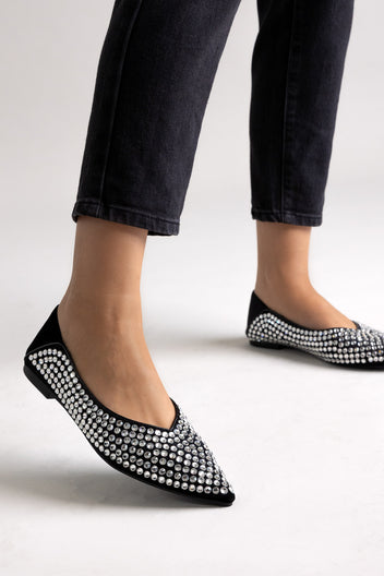 Black suede pointed-toe flats with rhinestone embellishment, angled view on a model in black jeans