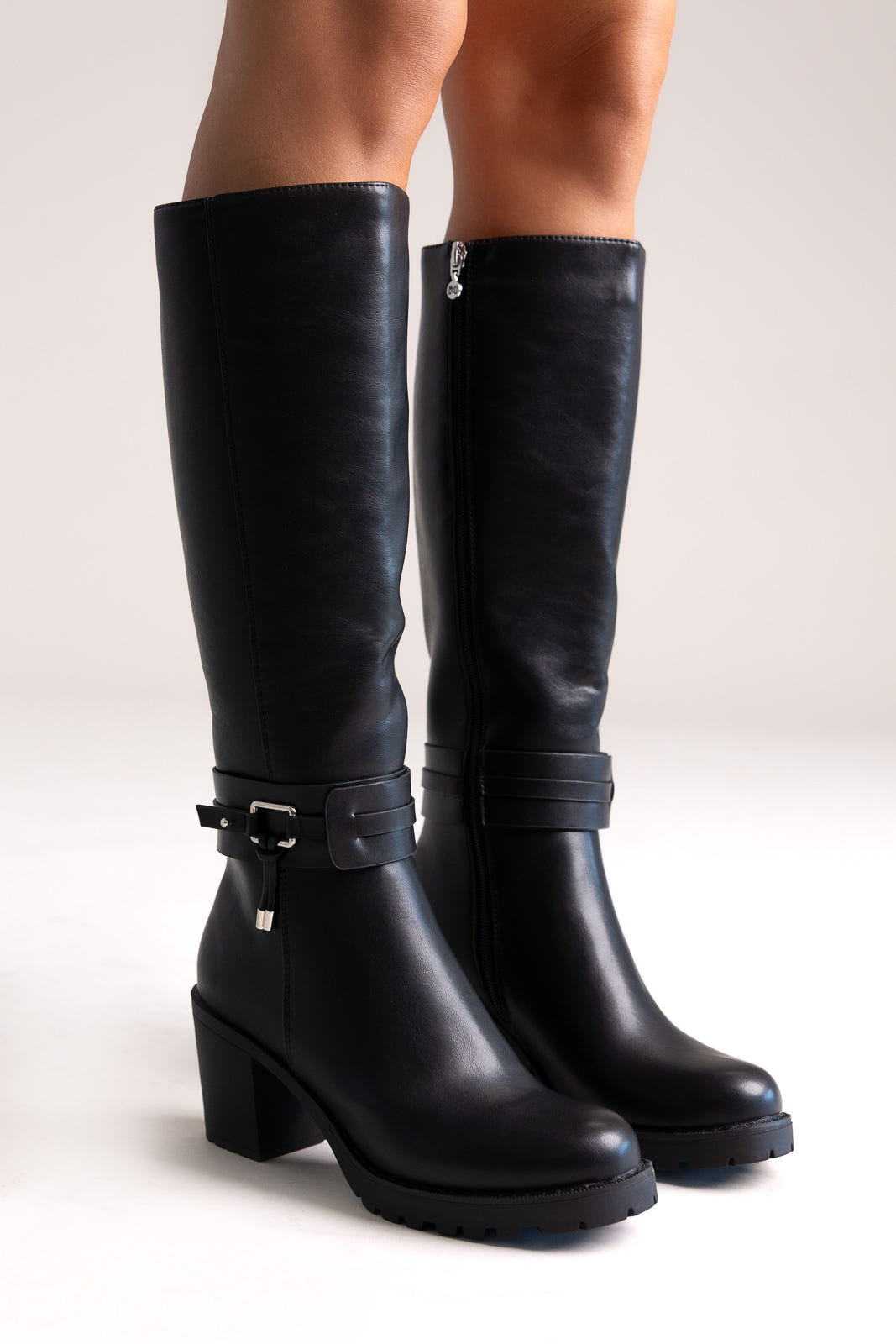 Model legs in black leather knee-high block heel boots with buckle strap and side zip, angled view on a light background