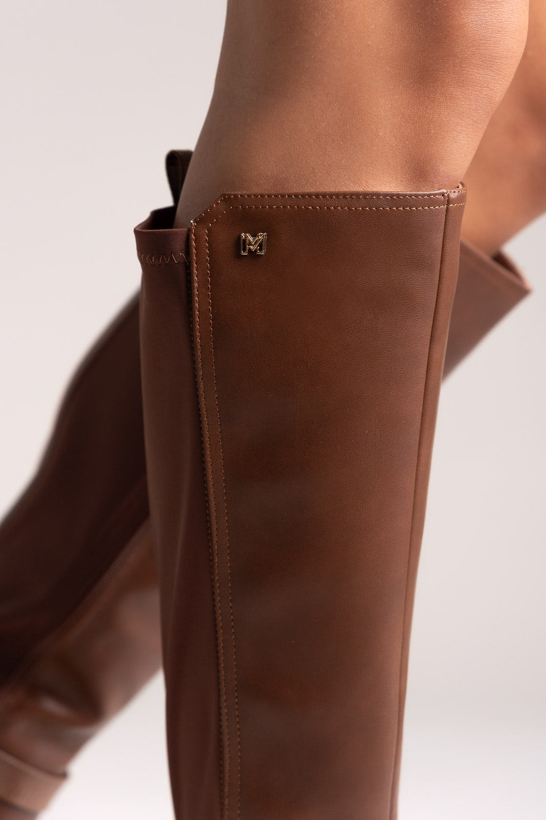 Close-up of tan brown leather knee-high boot shaft with small gold hardware at the top edge on a light background