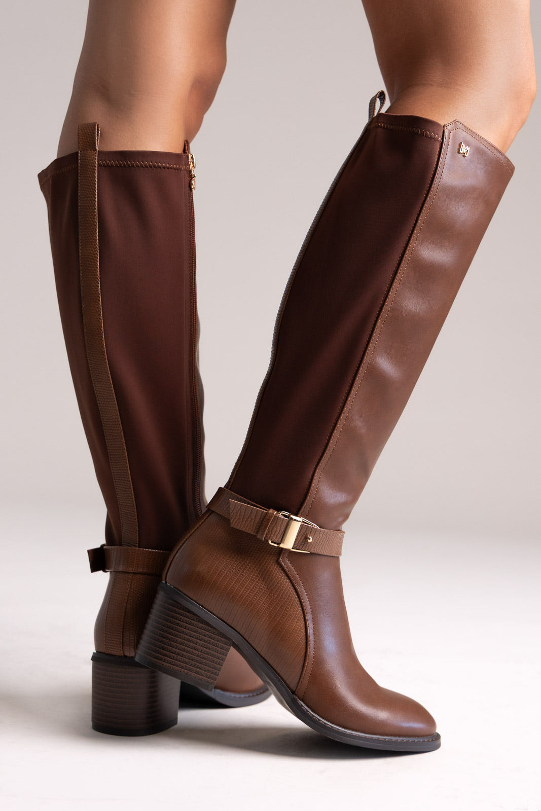 Model legs in tan brown knee-high block heel boots with buckle strap and stretch panel, angled view on a light background