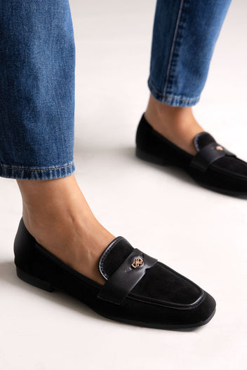 Black suede loafers with leather trim and gold-tone bit, side view on a model in grey trousers