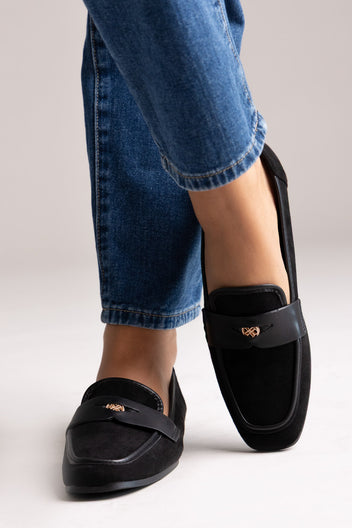 Black suede loafers with leather trim and gold-tone bit, angled view on a model in grey trousers