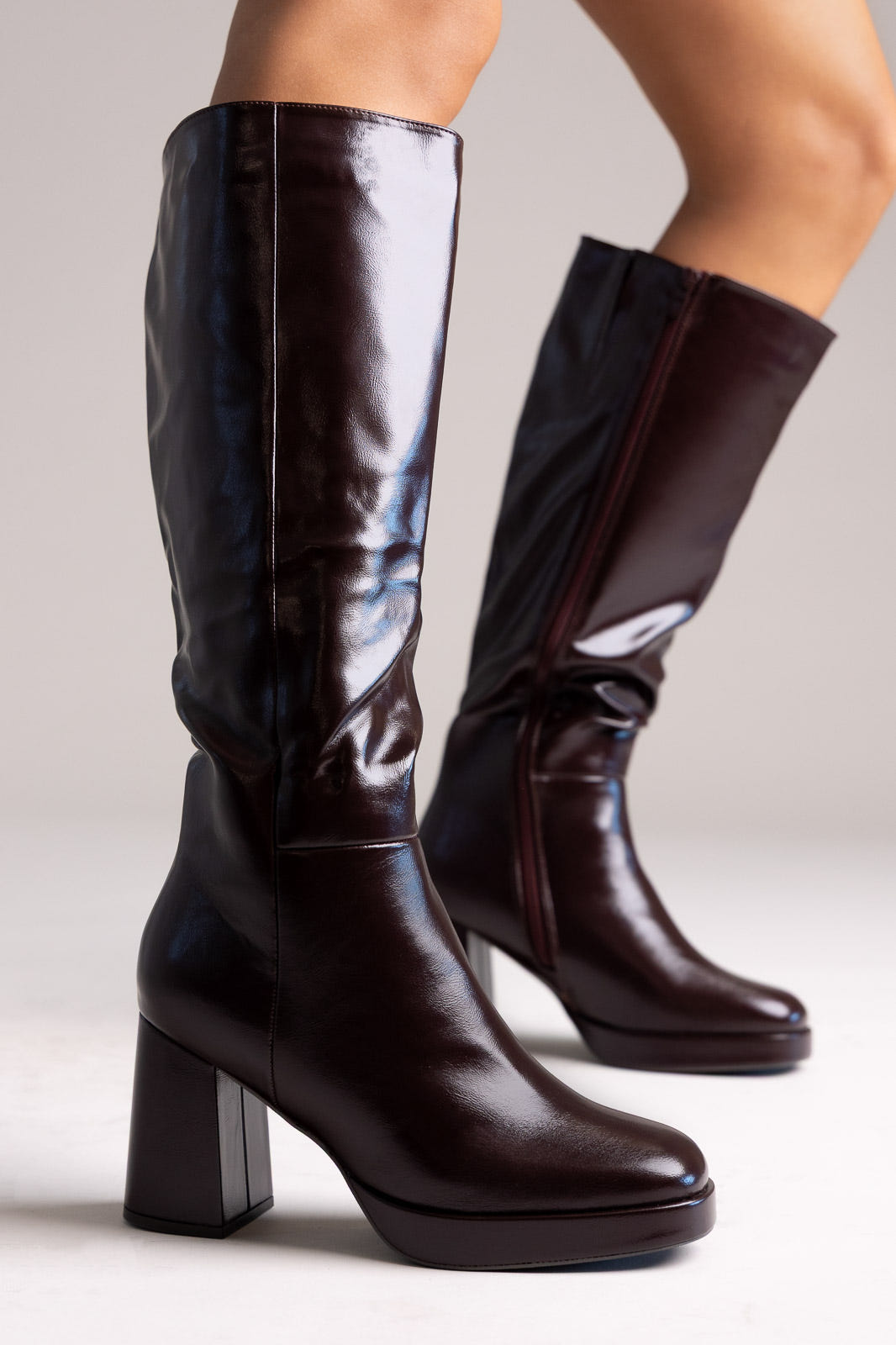 Dark burgundy glossy faux leather knee-high boots with platform and block heel, side view on a model