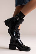 Black crinkled patent lace-up combat boots with buckle strap and lug sole, side view on a model