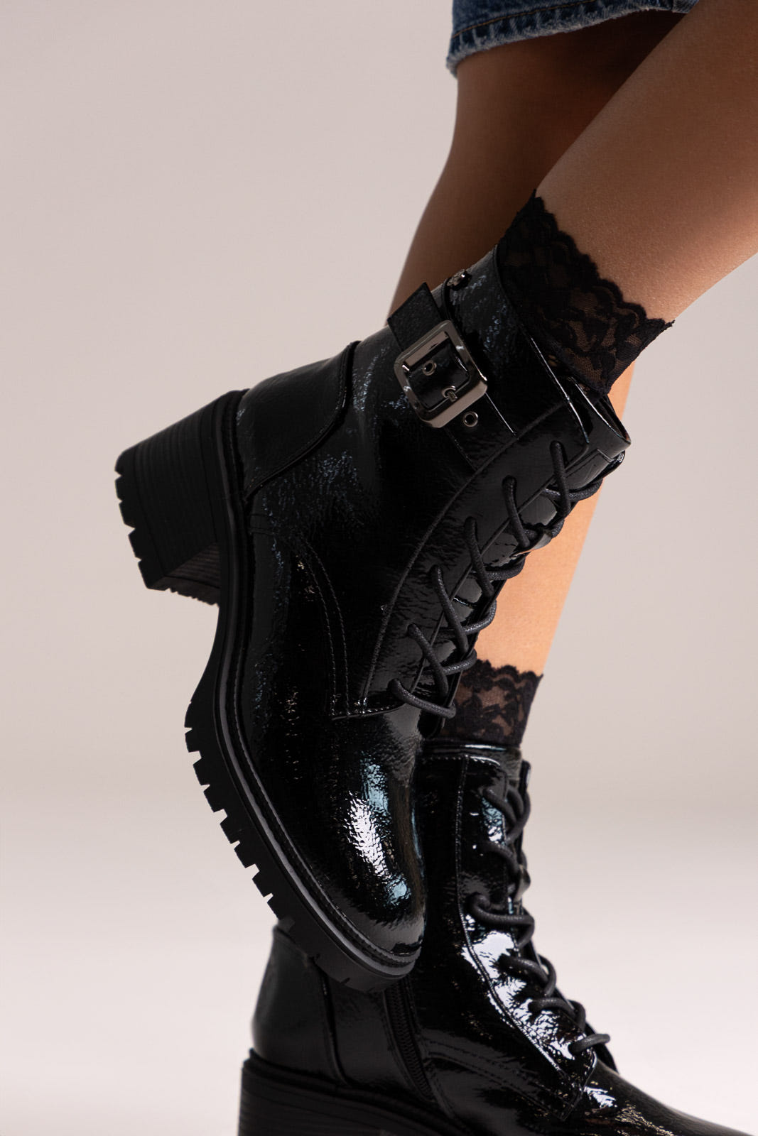 Black crinkled patent lace-up combat boots with buckle strap and lug sole, worn with lace socks on a model