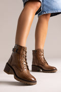 Model leg in brown leather lace-up ankle boot with buckle straps and block heel, side view on a light backdrop