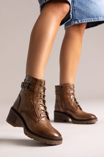 Model leg in brown leather lace-up ankle boot with buckle straps and block heel, side view on a light backdrop