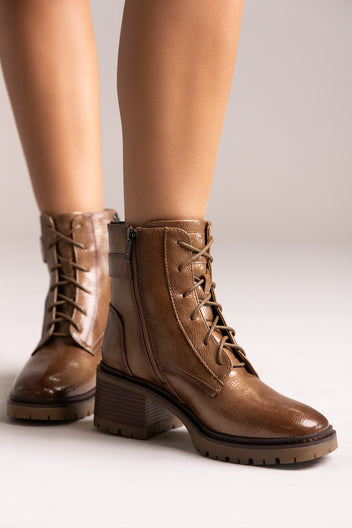 Model legs in brown leather lace-up ankle boots with buckle straps and block heels, angled view on a light backdrop