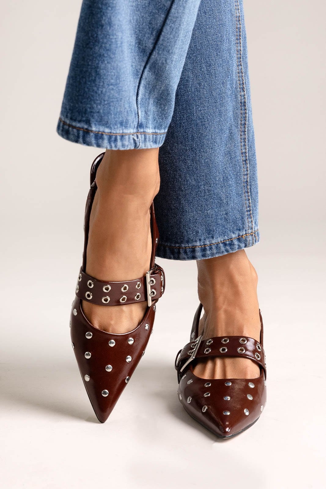 Burgundy leather pointed-toe slingback heels with studs and buckle strap, angled view on a model in jeans