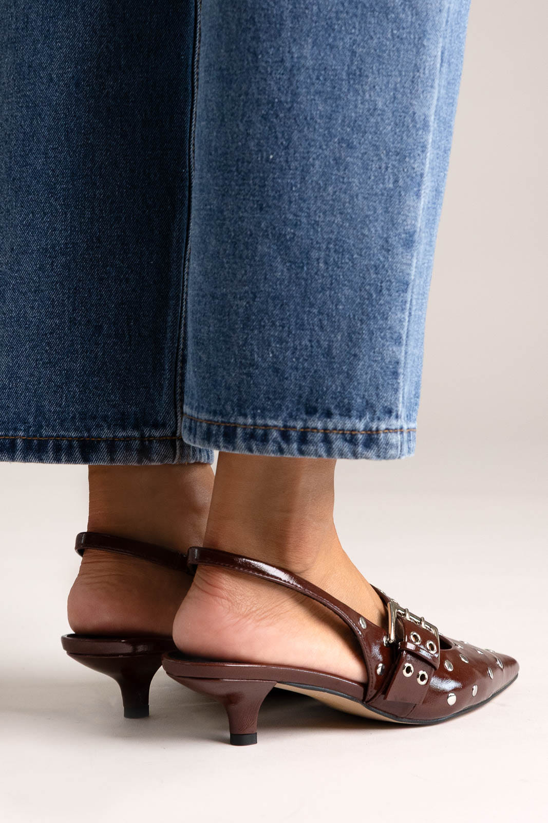 Burgundy leather pointed-toe slingback heels with grommets and buckle strap, side view on a model in jeans