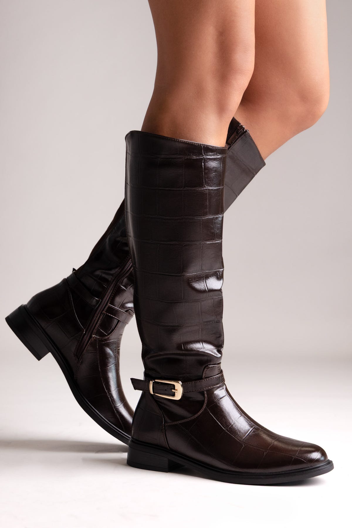 Dark brown croc-embossed knee-high riding boots with gold buckle strap and flat sole, side view on a model