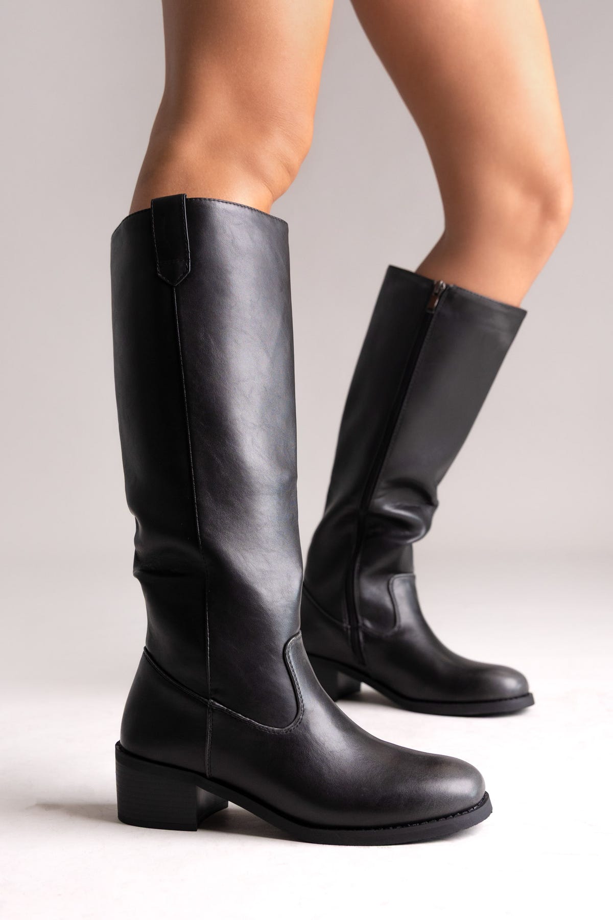 Model legs in black leather knee-high boots with round toes, chunky block heels and side zip, angled view on light background