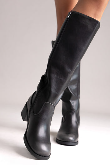 Model leg raised in black leather knee-high boot with smooth shaft and chunky block heel, angled view on a light background