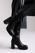 Model legs in black knee-high block heel boot with lug sole and side zip, angled view on light grey background