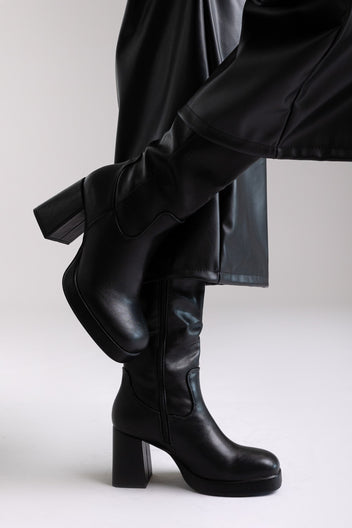 Model legs in black knee-high block heel boot with lug sole and side zip, angled view on light grey background
