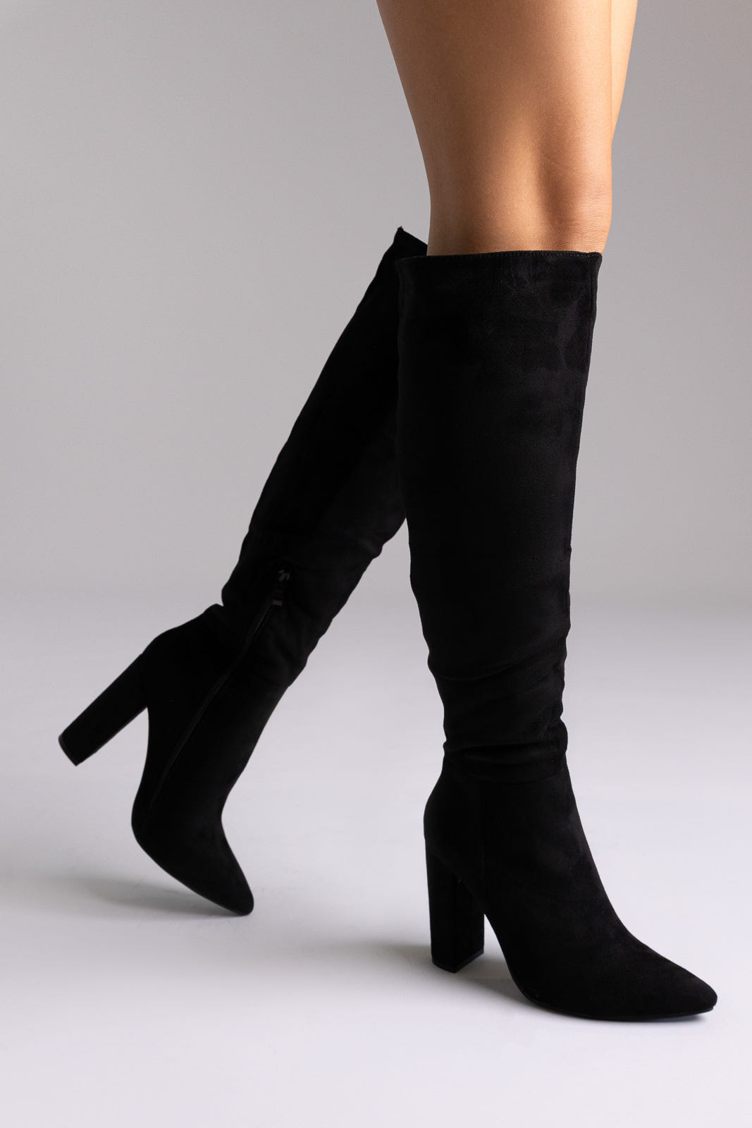Black suede over-the-knee boots with pointed toe and block heel, side view on white backdrop