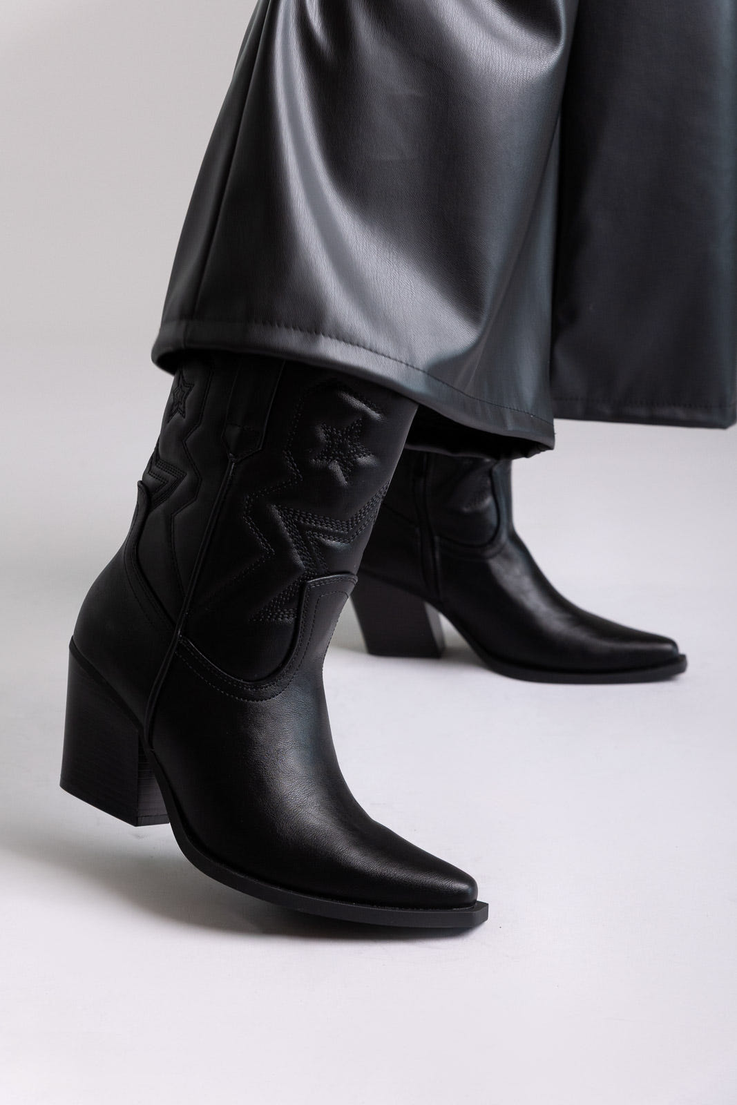 Black faux leather knee-high western boots with block heel and stitched shaft, angled view on a model