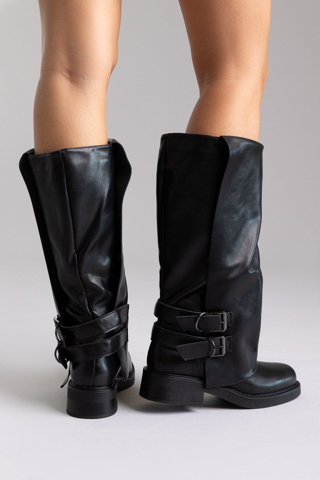 Model legs in black leather knee-high boots with buckle straps and round toes, front view on a light grey background