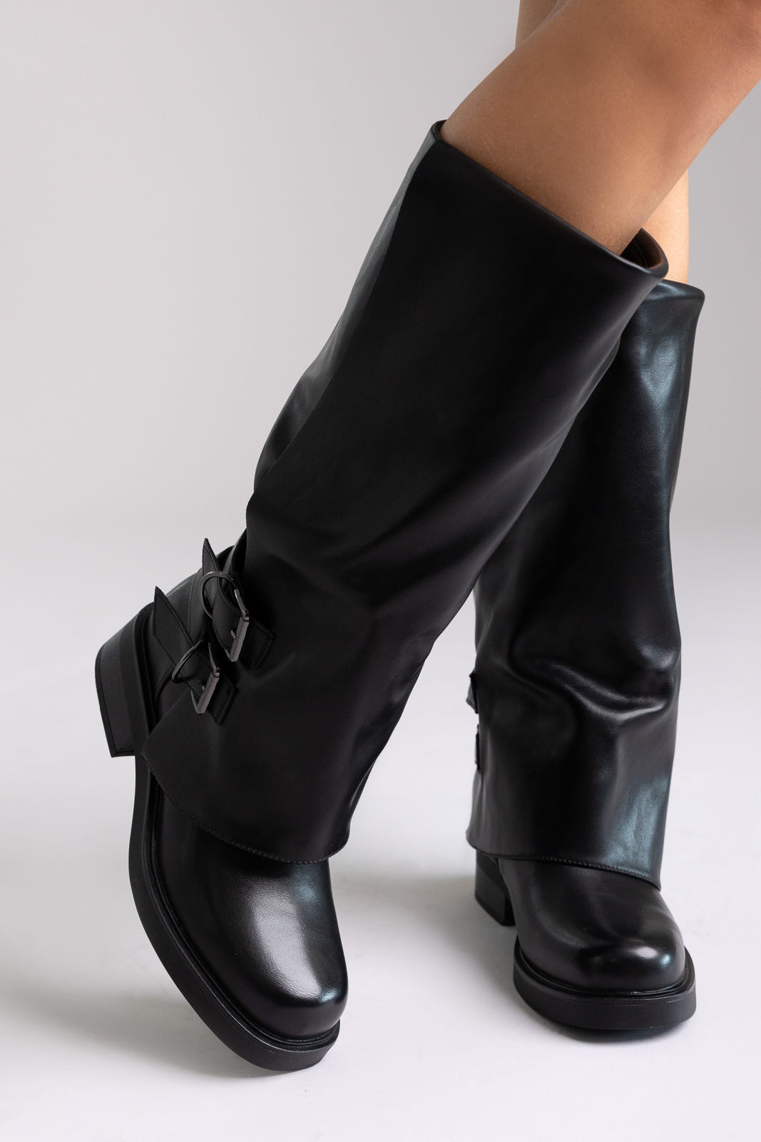 Model legs in black leather knee-high boots with buckle straps and chunky block heel, angled view on a light grey background