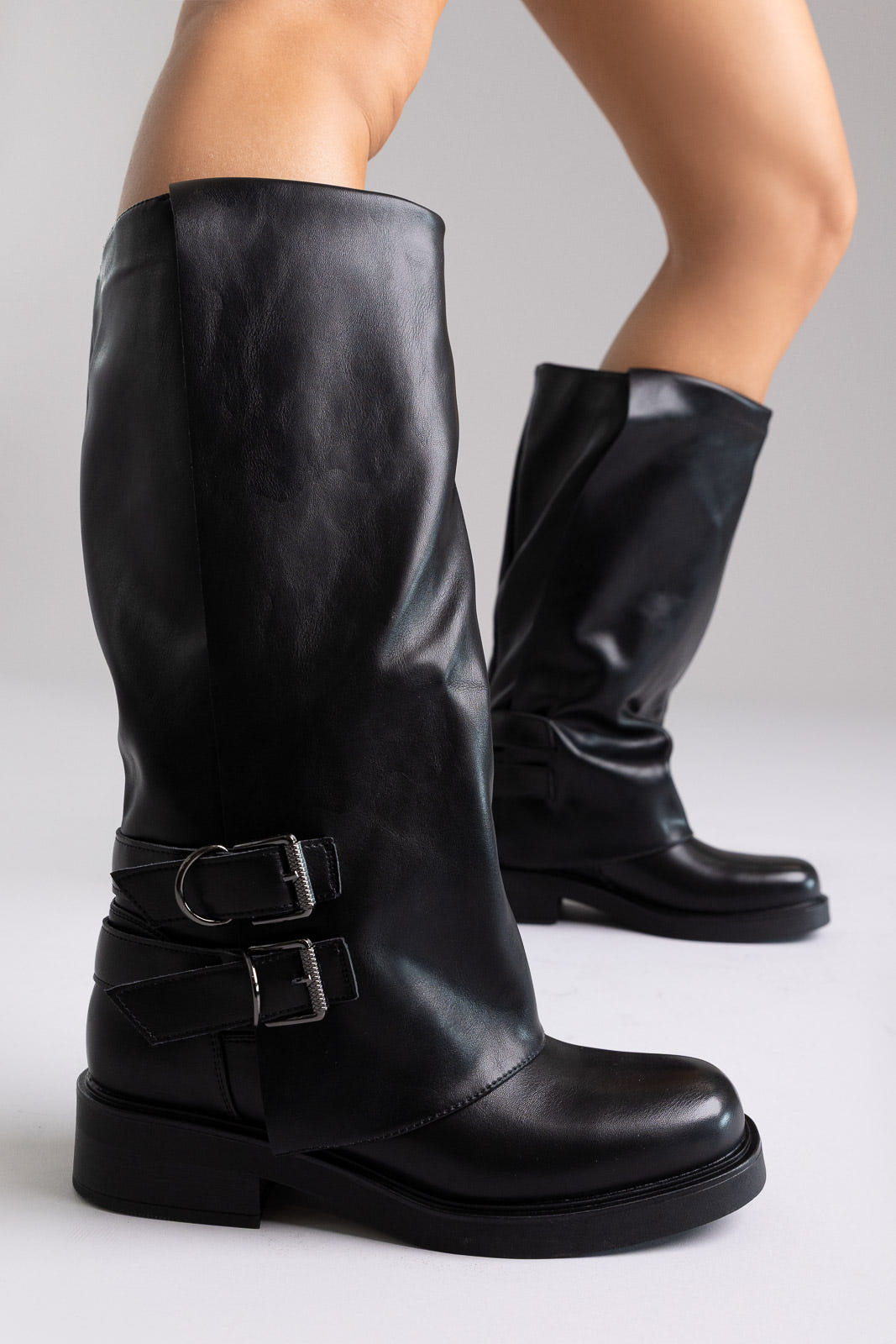 Pair of black leather knee-high boots with buckle straps and chunky sole, angled view on a light grey background