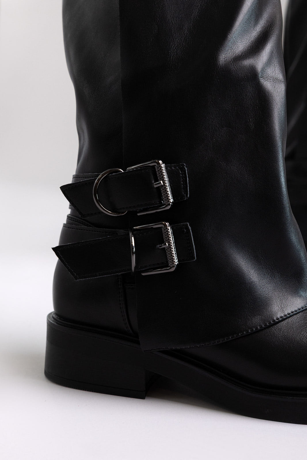 Close-up of black leather boot shaft with two buckle straps and a chunky sole on a light grey background
