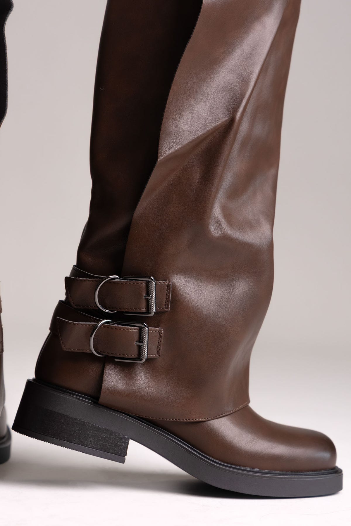 Brown leather knee-high boot with buckle straps and chunky sole, close side view on a light grey background